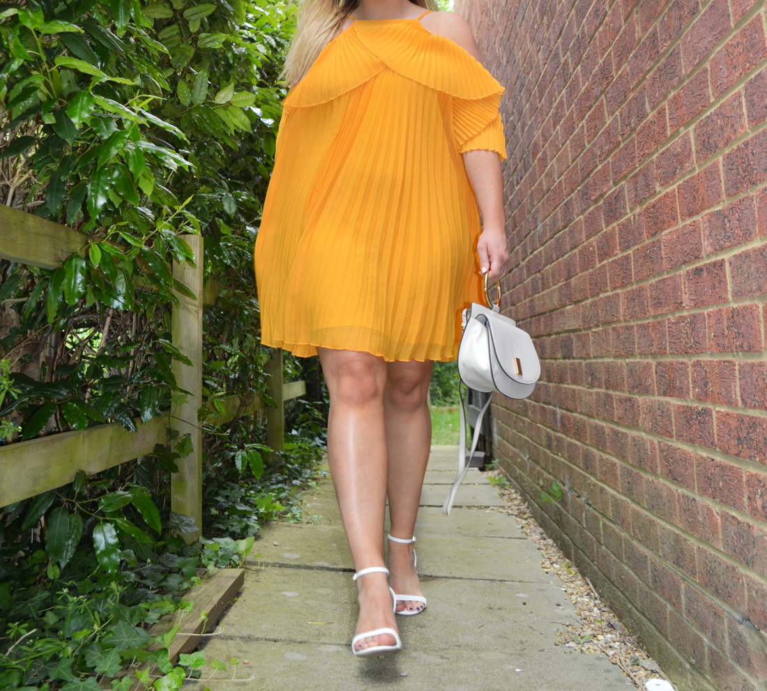 Hannah in a pleated and floaty yellow dress that comes just above the knee with barely there white heeled sandals with cream Mango bag with gold handle