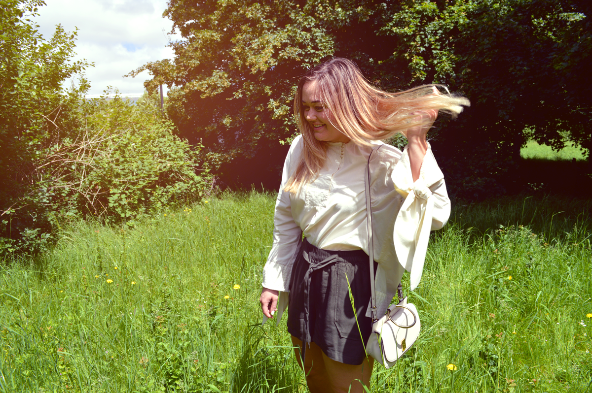 Hannah wearing Mango over-sized cream shirt with bows hanging down with khaki baggy shorts in a field on a sunny day.