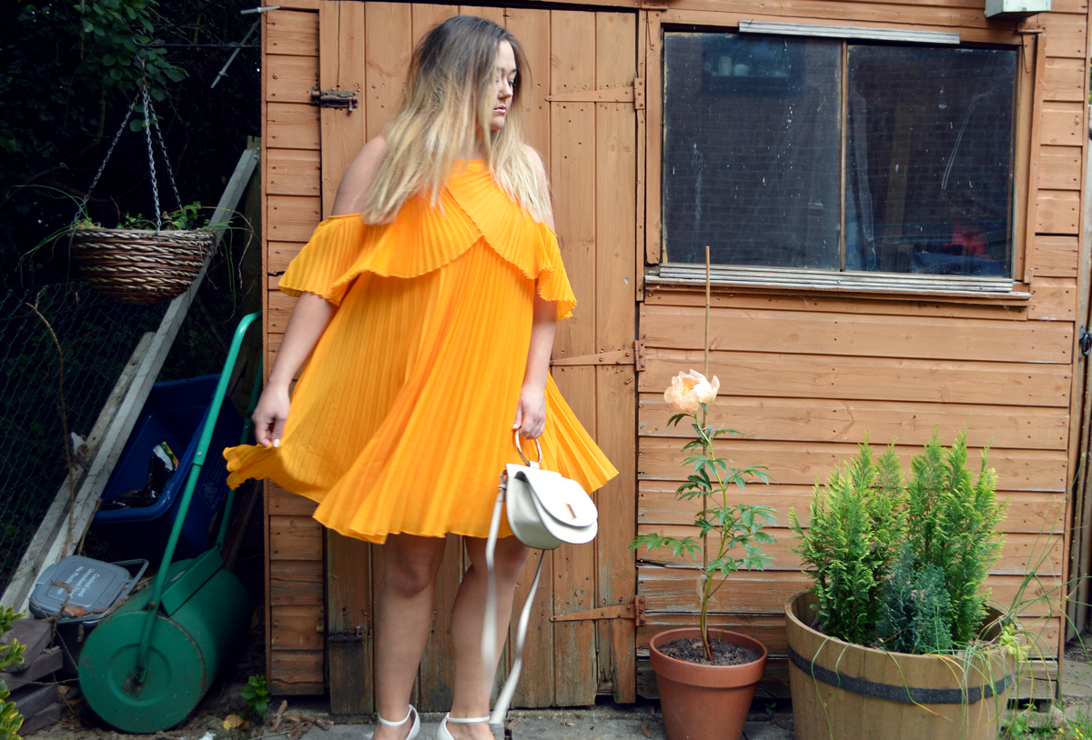 Hannah in a pleated and floaty yellow dress that comes just above the knee with barely there white heeled sandals with cream Mango bag with gold handle